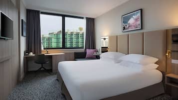 Ein Hotelzimmer mit einem großen Bett, einem Schreibtisch, einem Sessel, einem Fernseher und einem Fenster mit Blick auf Gebäude.