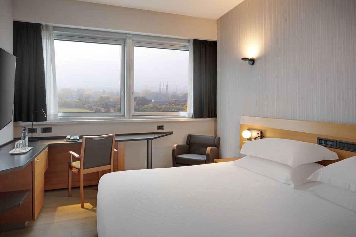 A hotel room with a bed, desk, and chairs. There is a window with a view of trees and buildings.