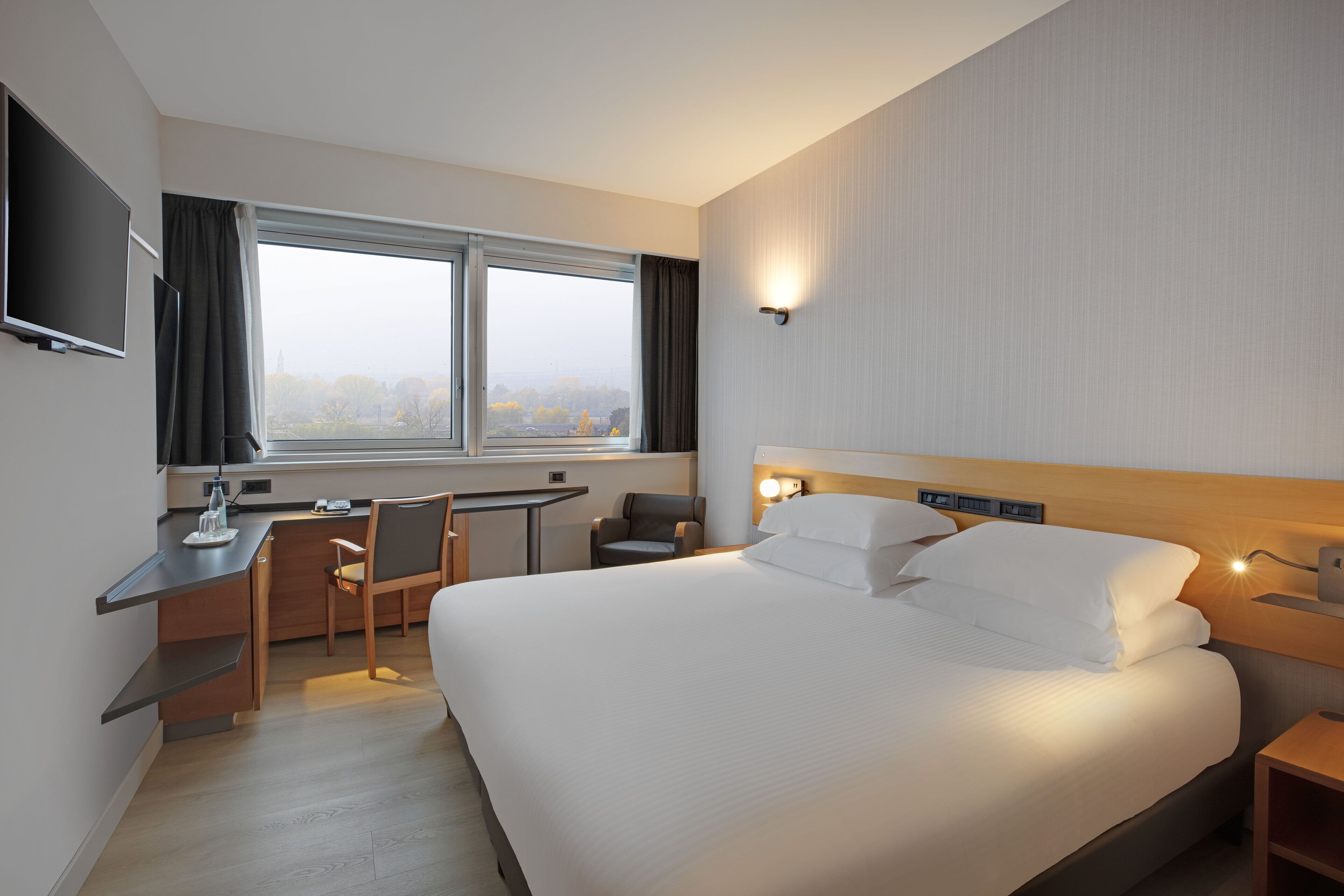 Et moderne hotelværelse med en stor seng, et skrivebord, en stol og et fjernsyn på væggen.