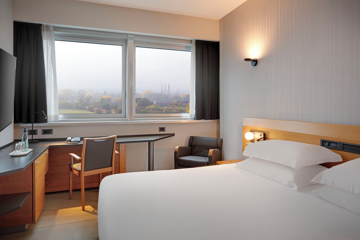 A hotel room with a bed, desk, chairs, a view through the window, and a nightstand with a lamp.