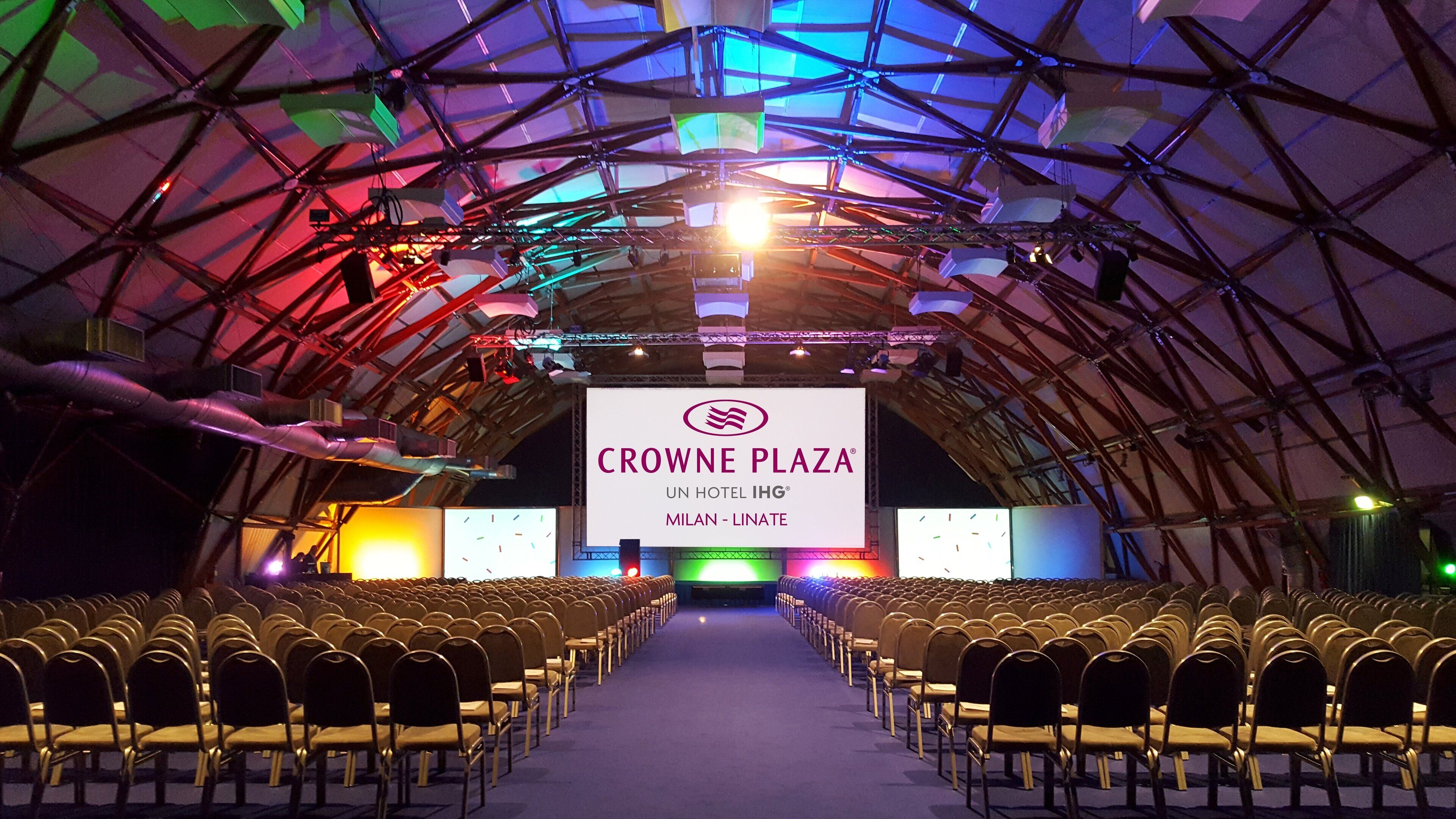 An indoor venue with rows of chairs facing a stage with a large screen displaying "CROWNE PLAZA UN HOTEL IHG MILAN - LINATE".