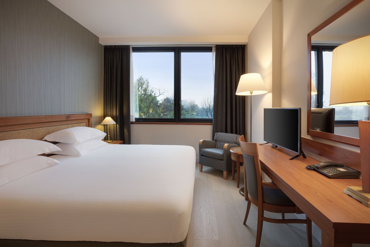 A hotel room with a large bed, a desk with a computer, a chair, and a window with curtains.