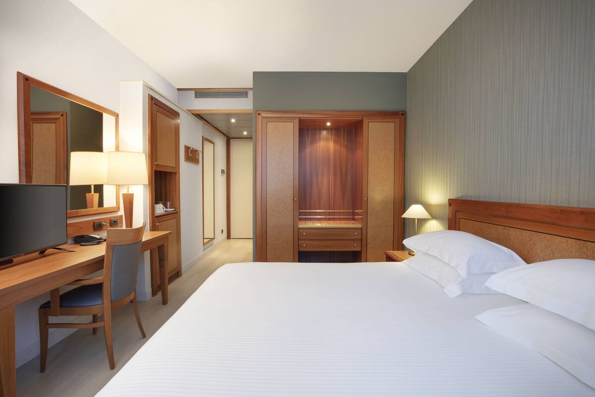 A hotel room with a bed, a desk with a chair, a television, a wardrobe, and a mirror.