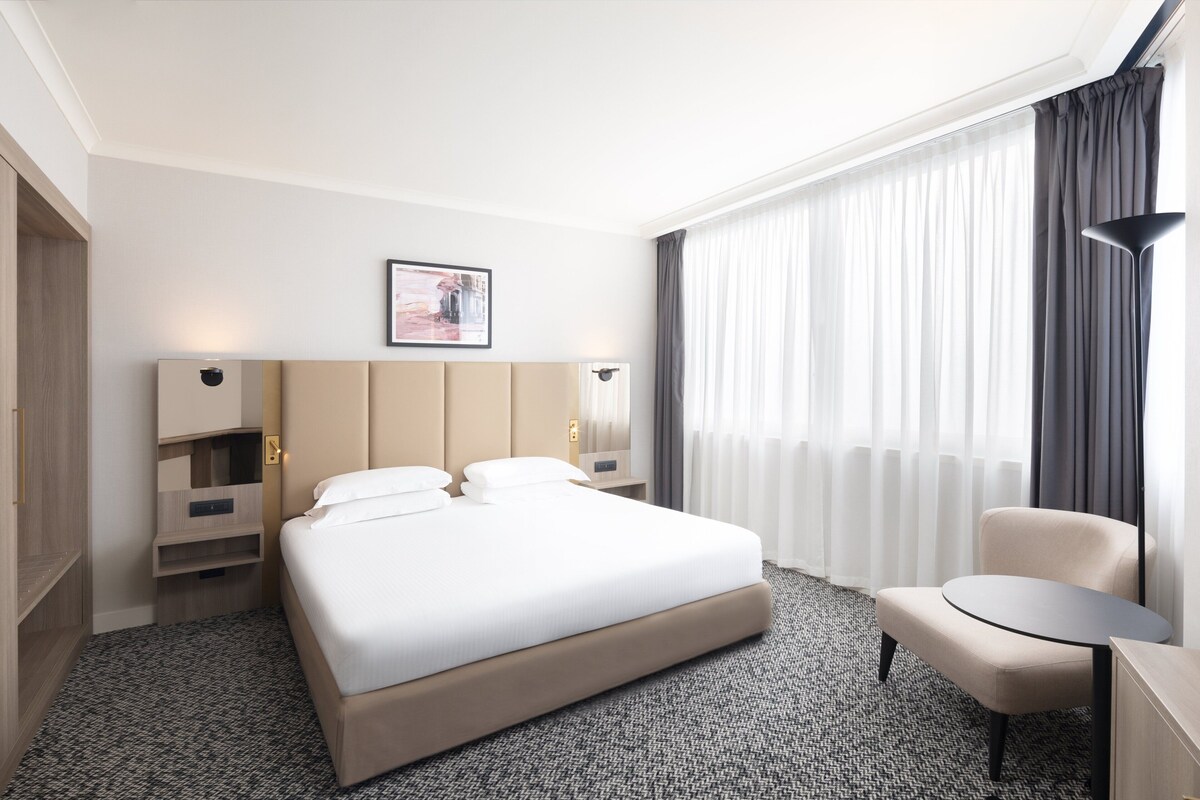 A hotel room with a large bed, bedside tables, a chair, and a large window with curtains.