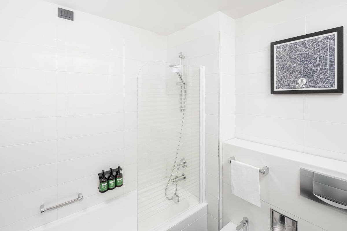 A modern bathroom with a glass-enclosed shower, white tiles, a framed map on the wall, and toiletries on the side.