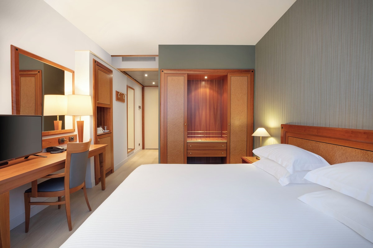 A hotel room with a bed, a desk with a chair, a television, and a wardrobe.