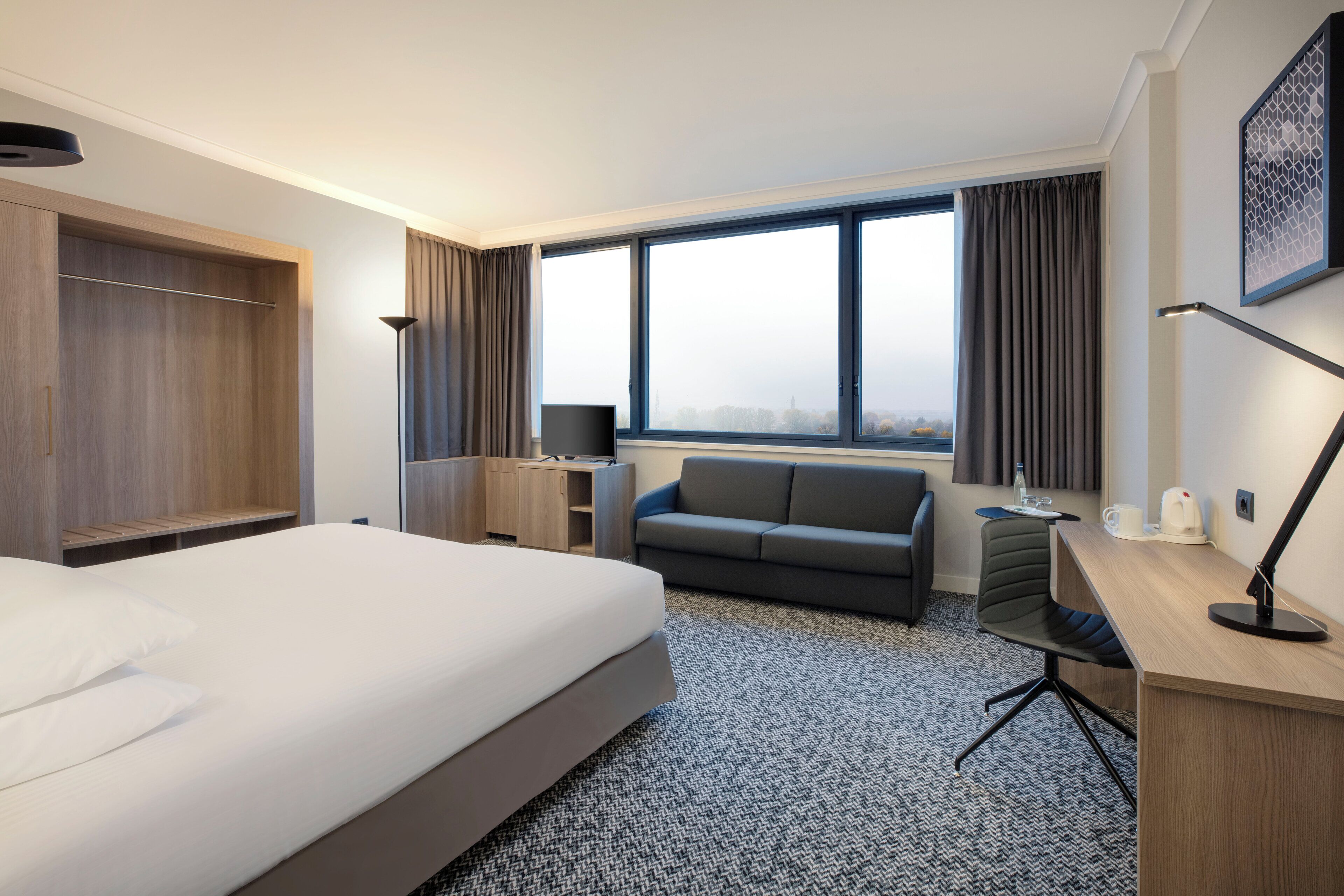 A hotel room with a bed, a sofa, a desk, a chair, a TV, and a window with curtains.