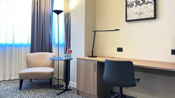 A hotel room with a desk, two chairs, a lamp, a small table with bottles, and a painting on the wall.