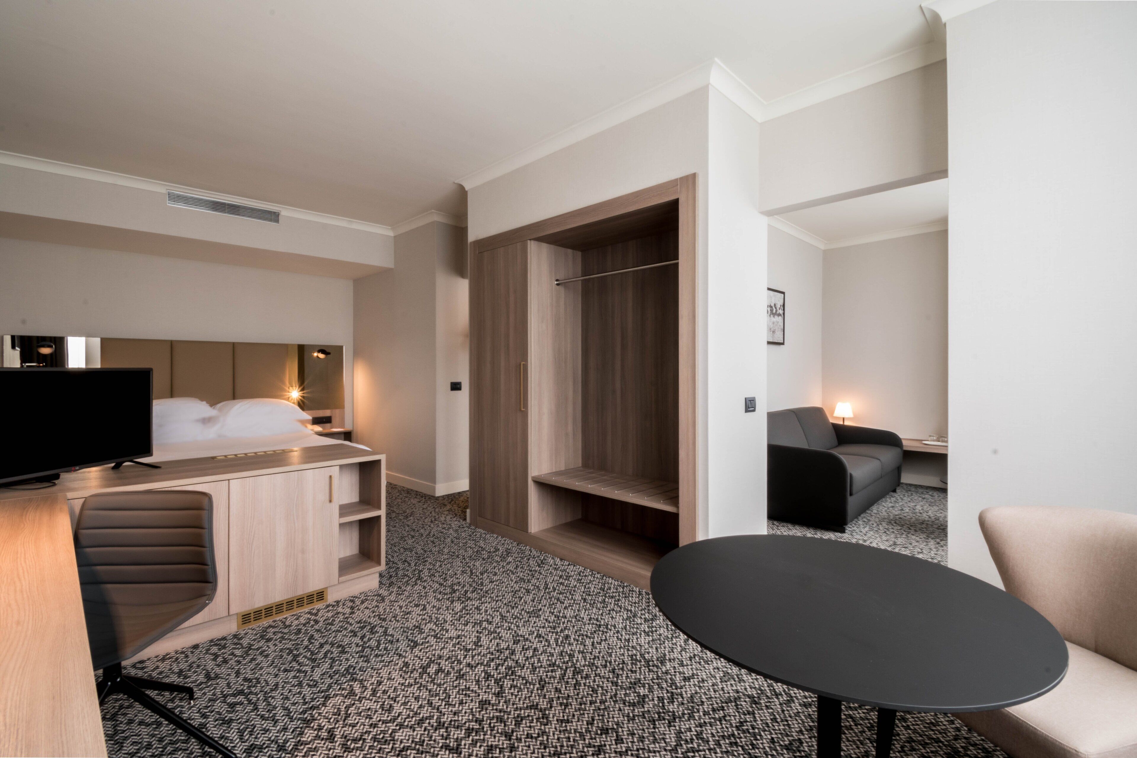 A hotel room with a bed, a desk with a TV, a wardrobe, a sofa, and a small table.