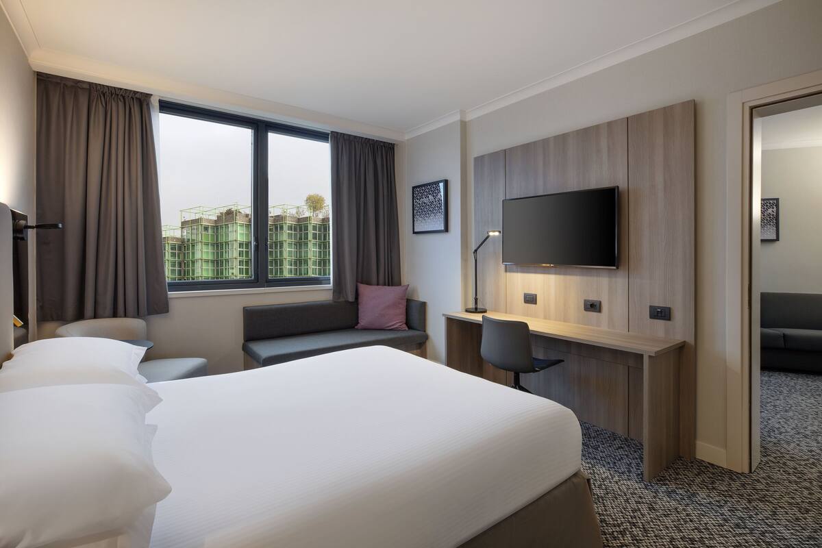 A hotel room with a large bed, a desk, a chair, a TV, and a window with curtains.