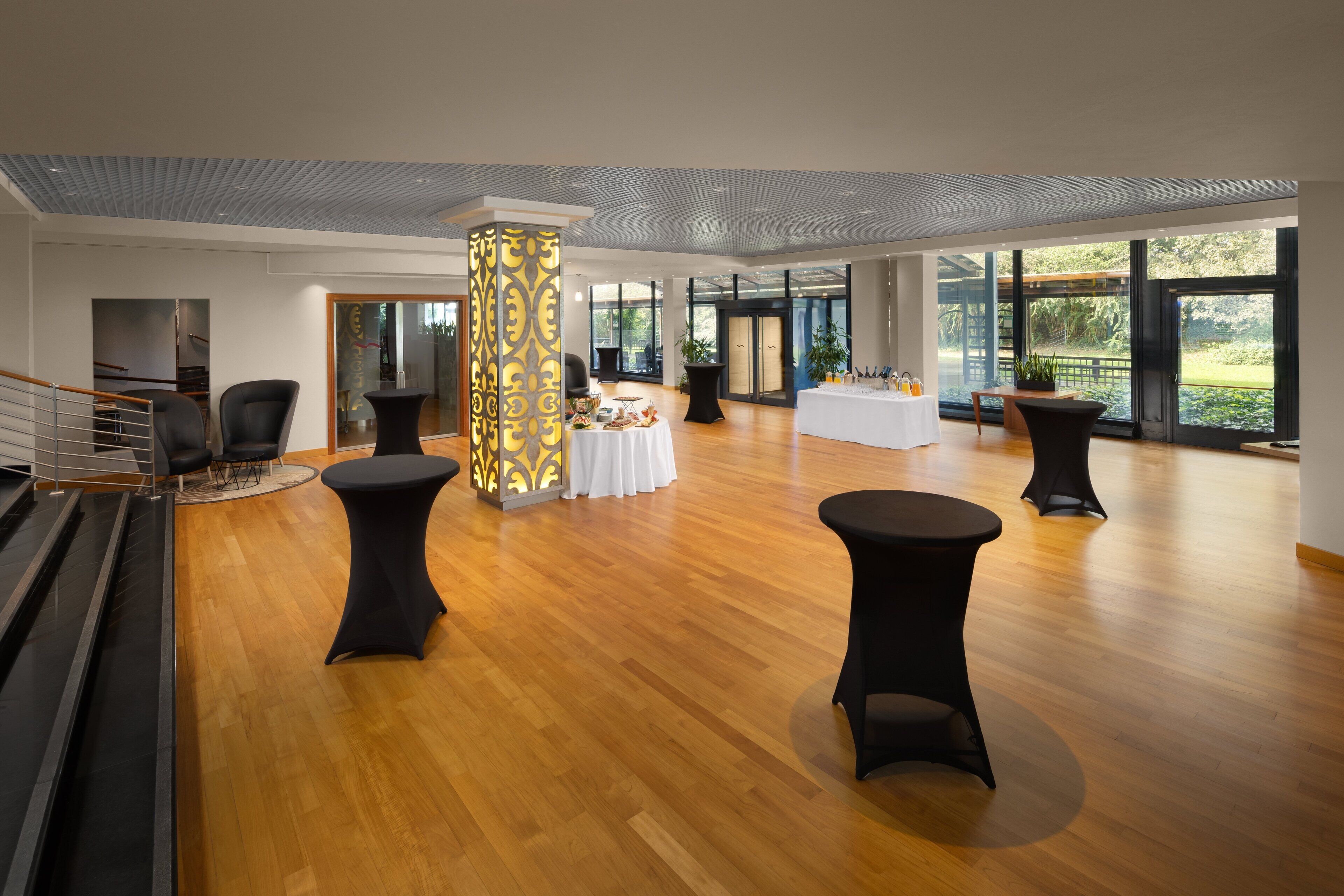 A spacious lobby with a wooden floor, black round tables, and a central decorative pillar.