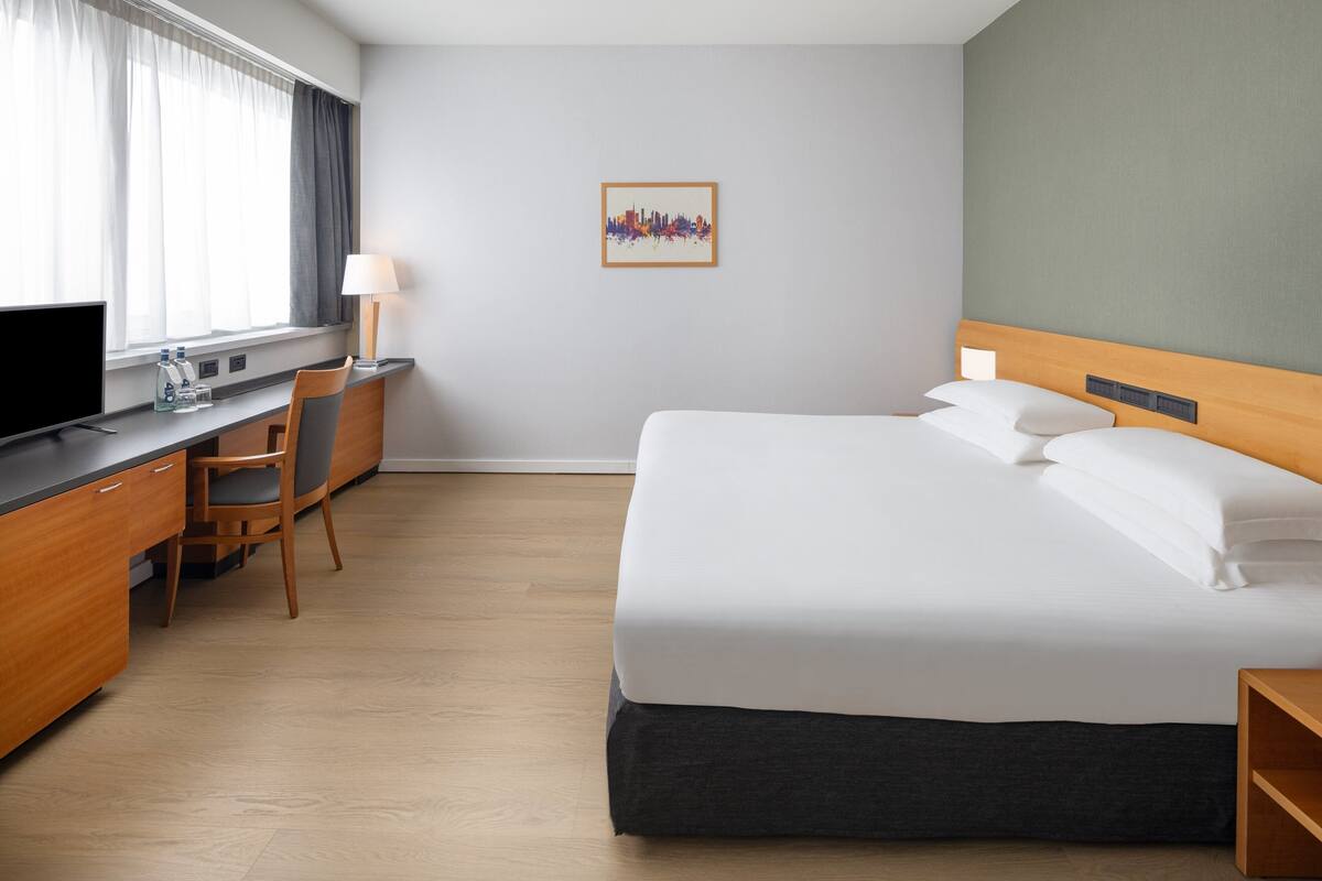 A hotel room with a bed, a desk, a chair, a TV, and a cityscape artwork on the wall.