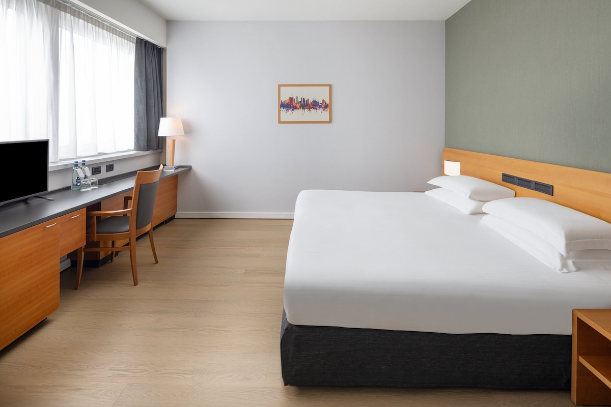 A hotel room with a bed, a desk, a chair, a TV, and a cityscape artwork on the wall.