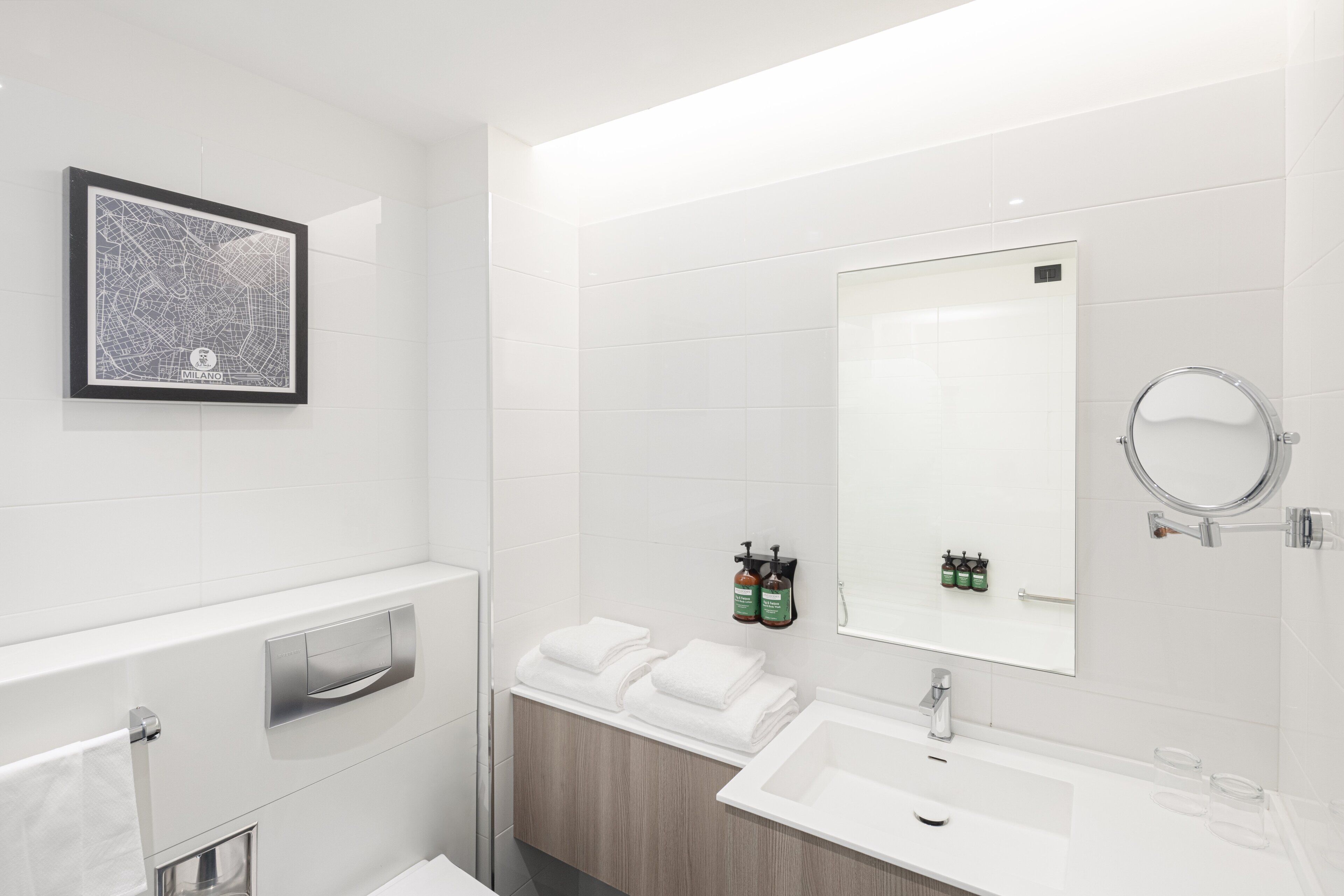 Une salle de bain moderne avec un grand miroir, des carreaux blancs, une carte encadrée et des serviettes soigneusement pliées.