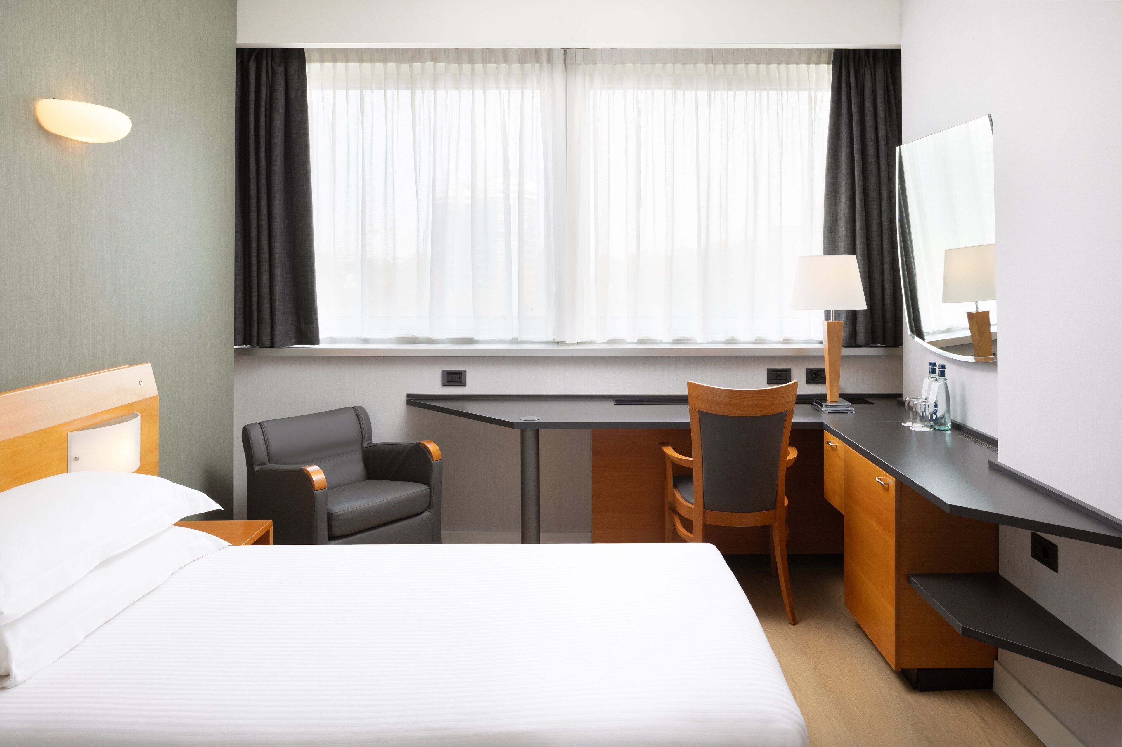 A hotel room with a bed, a desk, a chair, a lamp, a mirror, and a window with curtains.