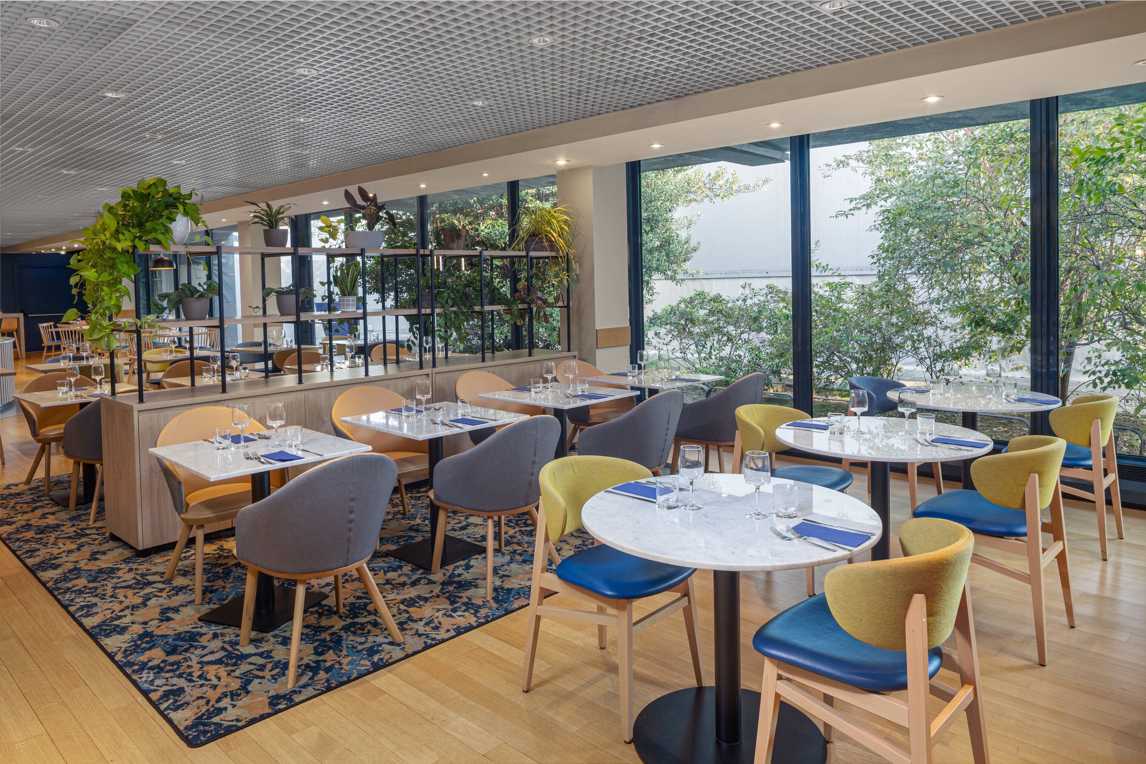 A modern restaurant with round tables and chairs, large windows, and indoor plants.