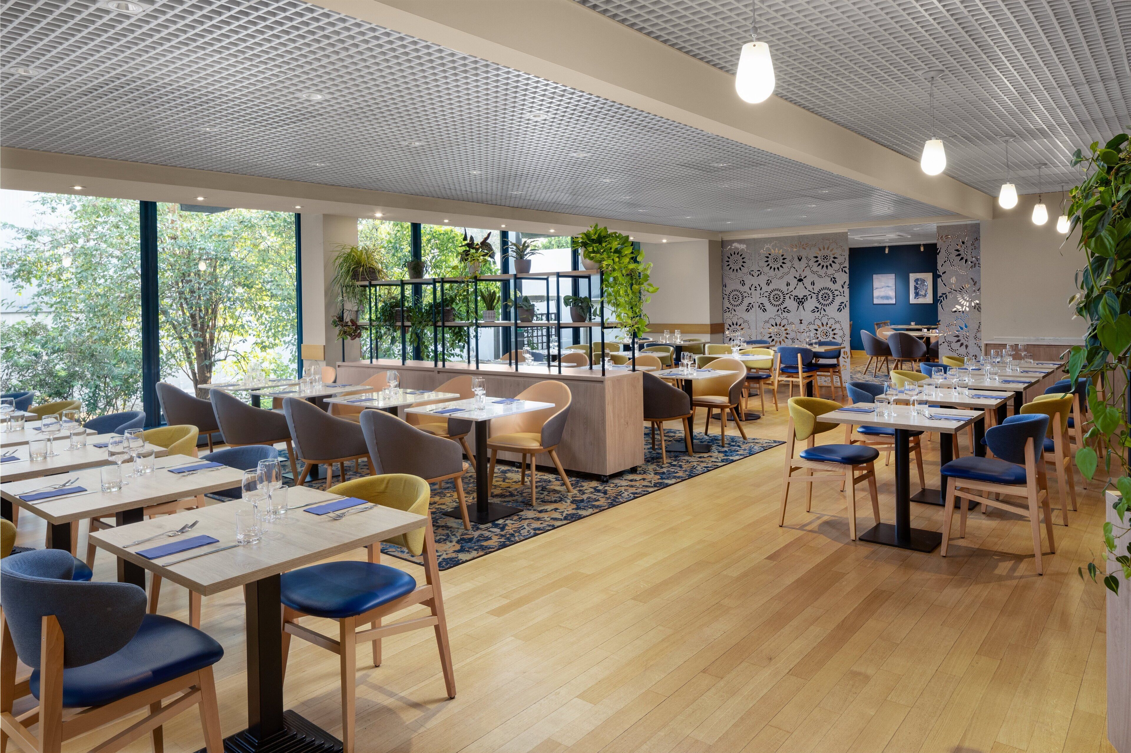 A modern dining area with multiple tables and chairs, large windows, and indoor plants.