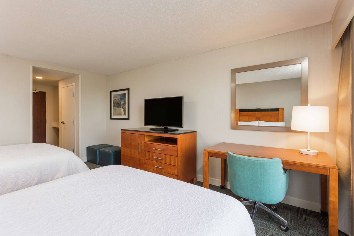 A hotel room with two beds, a wooden desk, a chair, a television, a dresser, and a mirror.