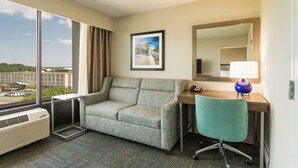 Habitación de hotel con un ventanal grande, un sofá, un escritorio con una silla, una lámpara y un cuadro en la pared.
