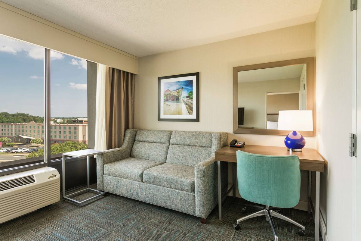 A hotel room with a large window, a sofa, a desk with a chair, a lamp, and a framed picture on the wall.
