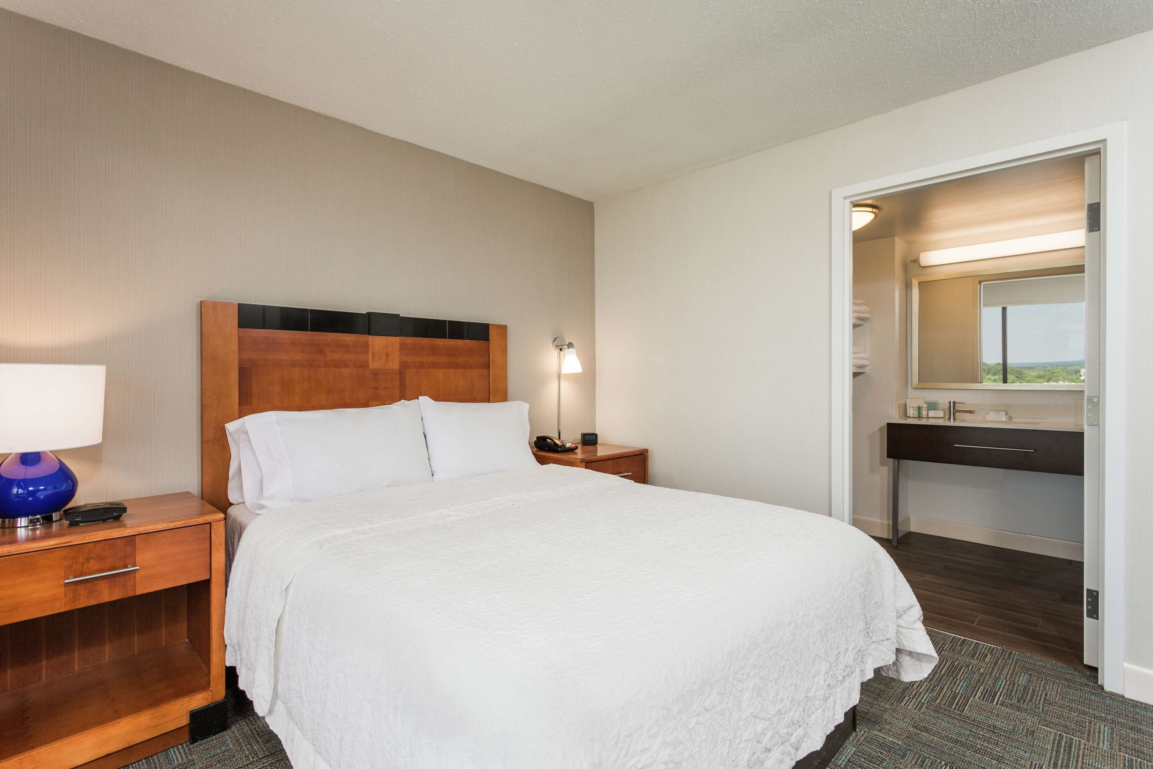Habitación de hotel con una cama grande, una mesita de noche de madera, un escritorio y un ba ño con espejo y lavamanos.