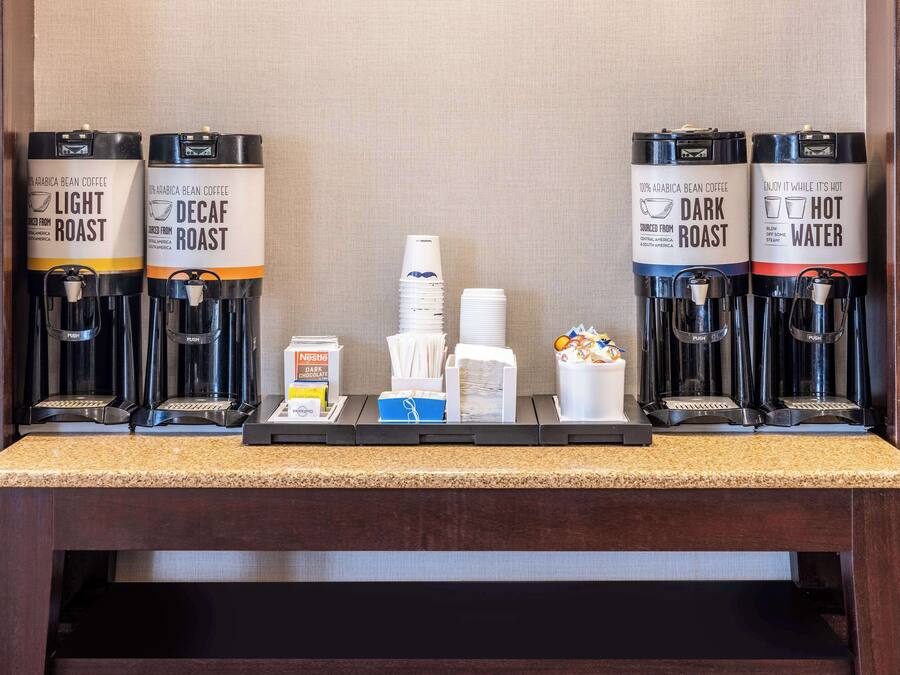 En kaffestation med fyra kaffedispensrar märkta med "Ljust rostad", "Koffeinfritt", "Mörkt rostad" och "Varmt vatten". Där finns staplar med engångsmuggar, en förpackning med servetter och en behållare med sockerbitar.