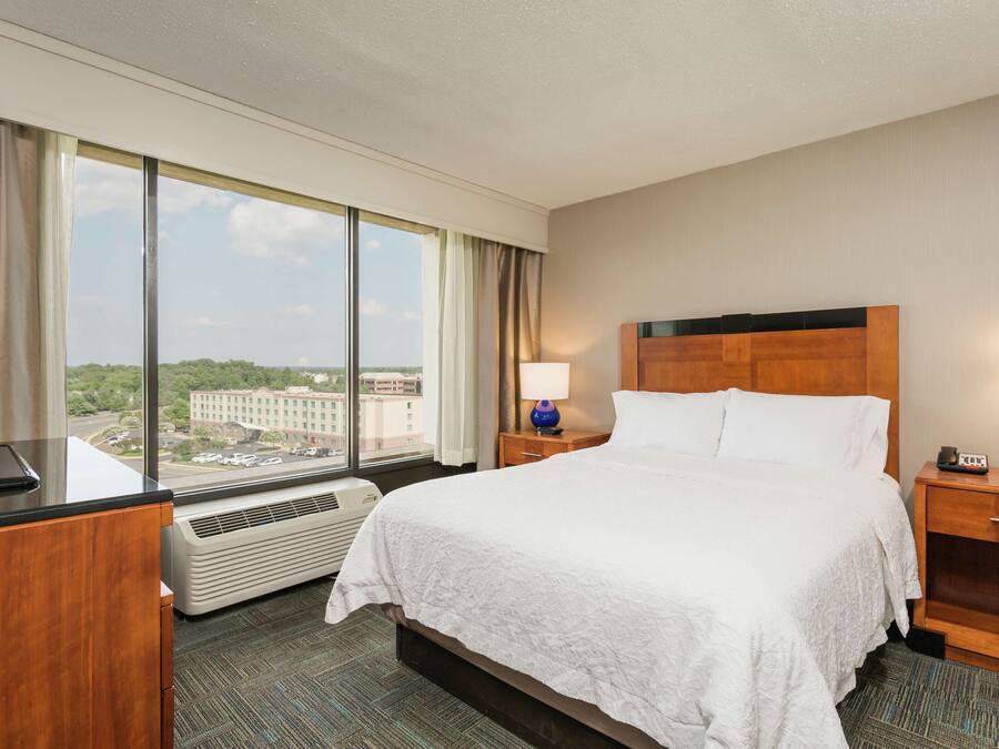 A hotel room with a large bed, a desk with a flat-screen TV, a nightstand with a lamp, and a view of a cityscape through large windows.