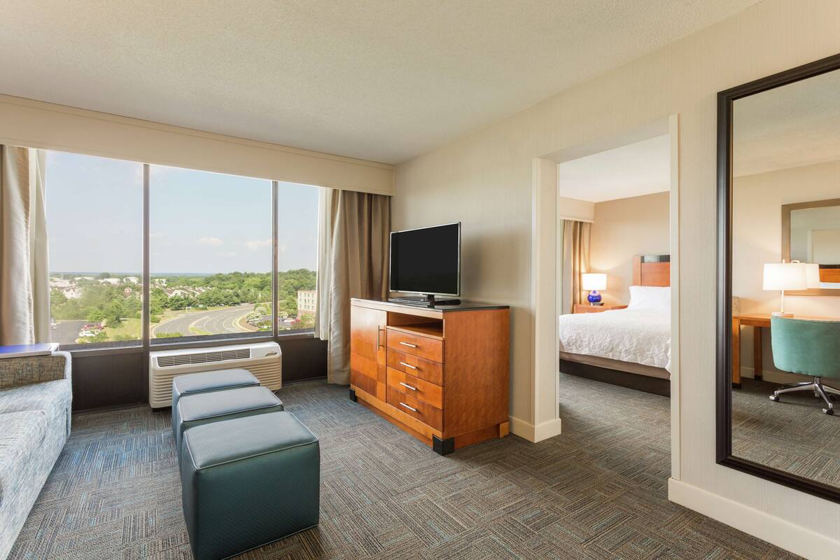 A hotel room with a large window, a flat-screen TV, a bed, a desk, and a chair.