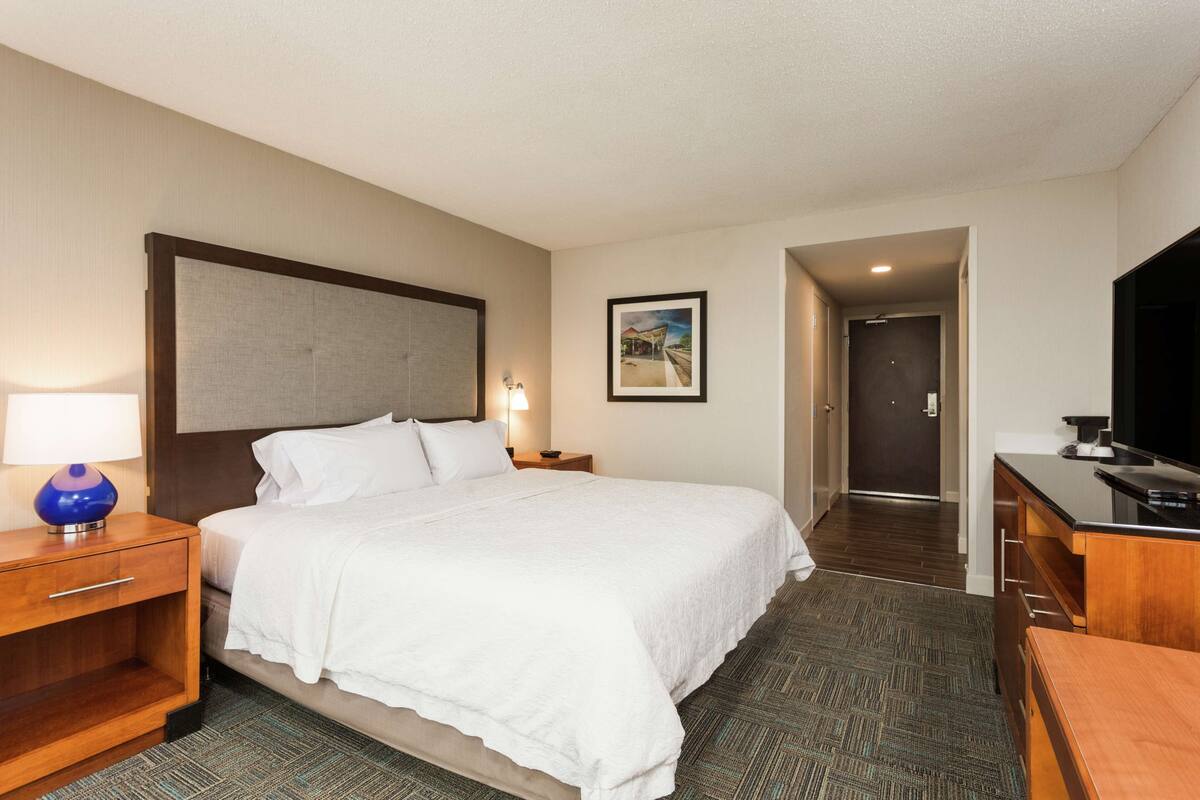 A hotel room with a bed, bedside table, lamp, picture on the wall, and a TV.