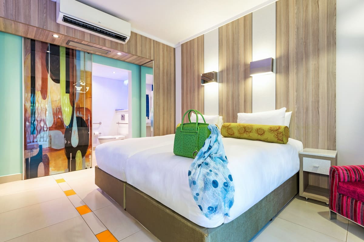 Une chambre d’hôtel avec un lit, une table de chevet, un sac à main vert et un tableau abstrait aux couleurs vives accroché au mur.