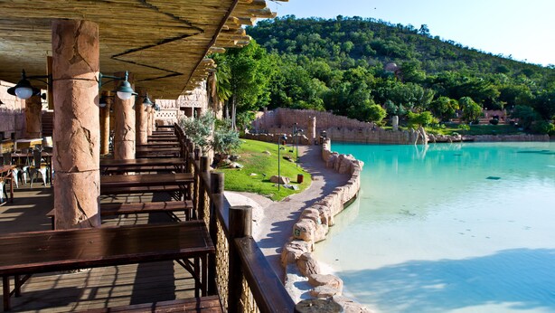 Un restaurant situé au bord de la rivière, doté de piliers en pierre, de bancs en bois et offrant une vue sur une rivière turquoise et des collines verdoyantes.