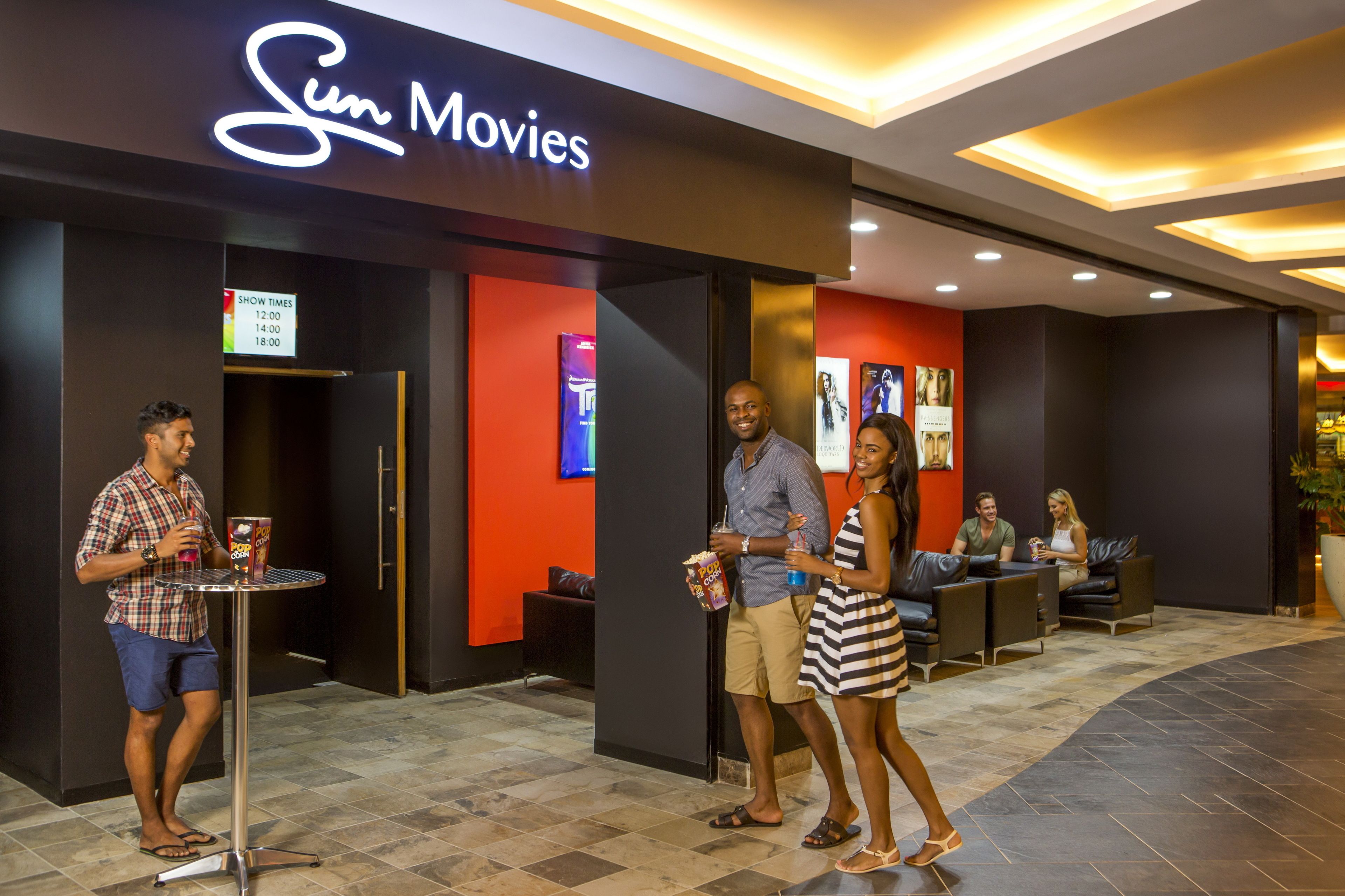 A cinema with a neon sign, a seating area with tables and chairs, and a signboard displaying show times.