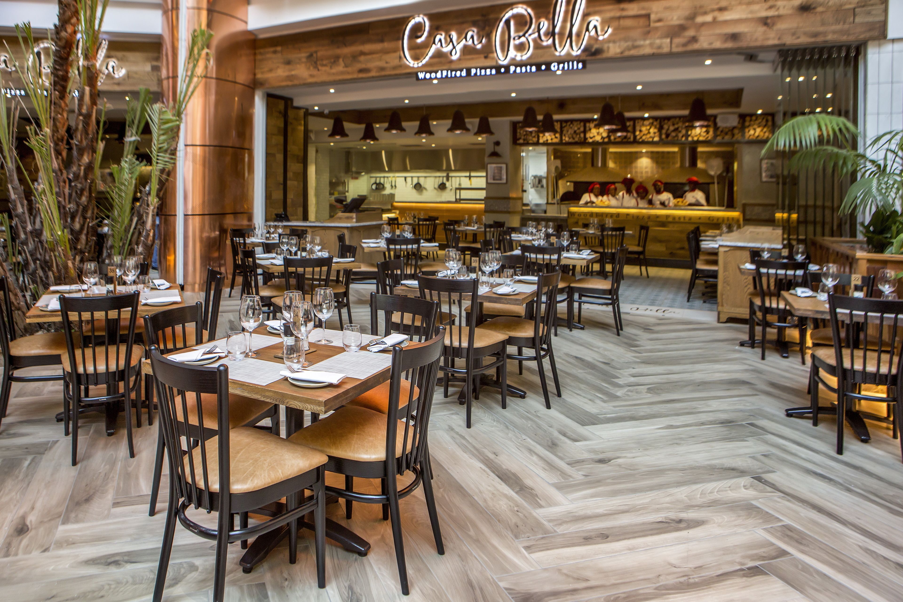 A restaurant with multiple tables and chairs, a wooden floor, and a visible kitchen area.