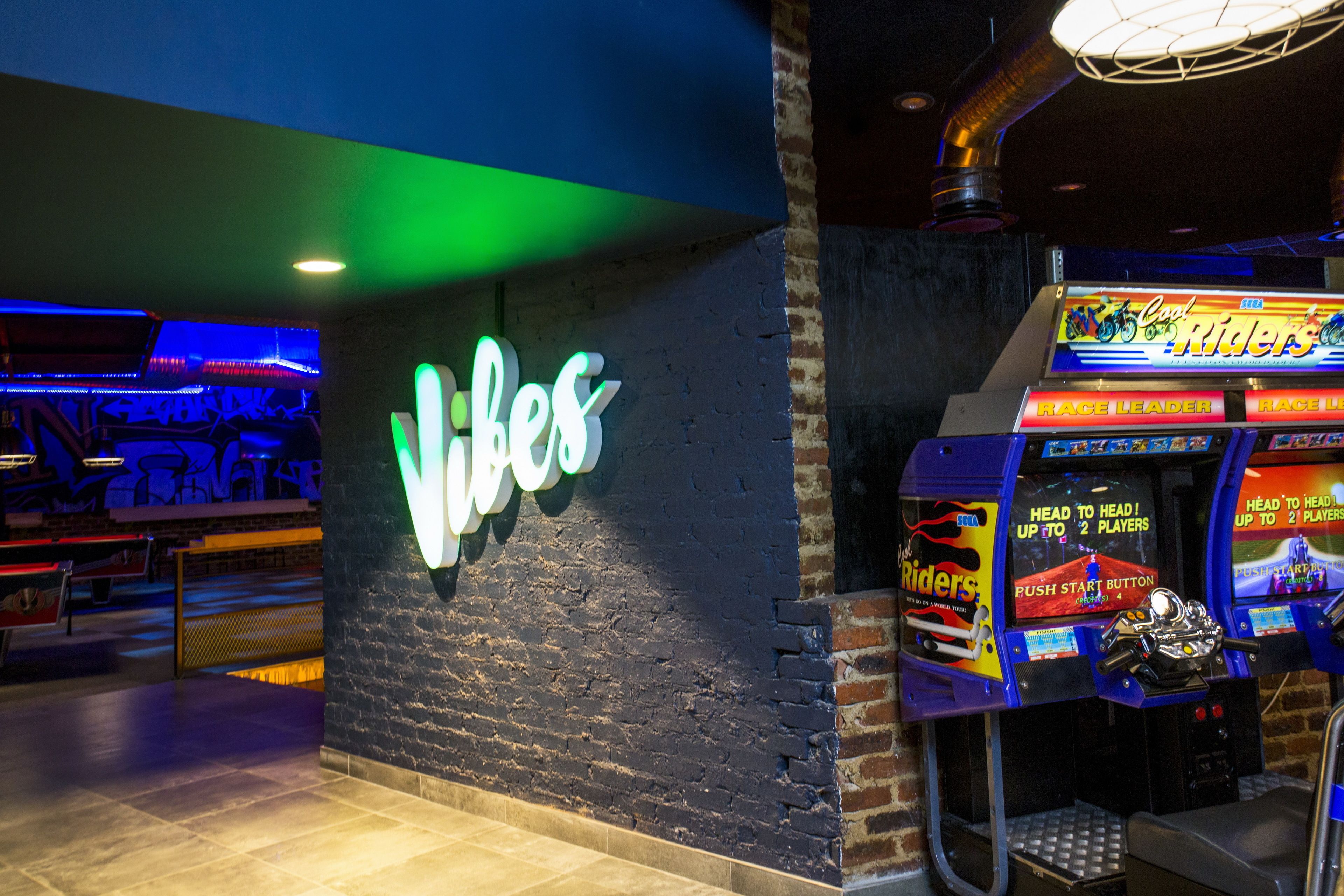 Un letrero de neón que dice "Vibes" sobre un muro de ladrillo, máquinas de videojuegos y una zona de estar con mesas.