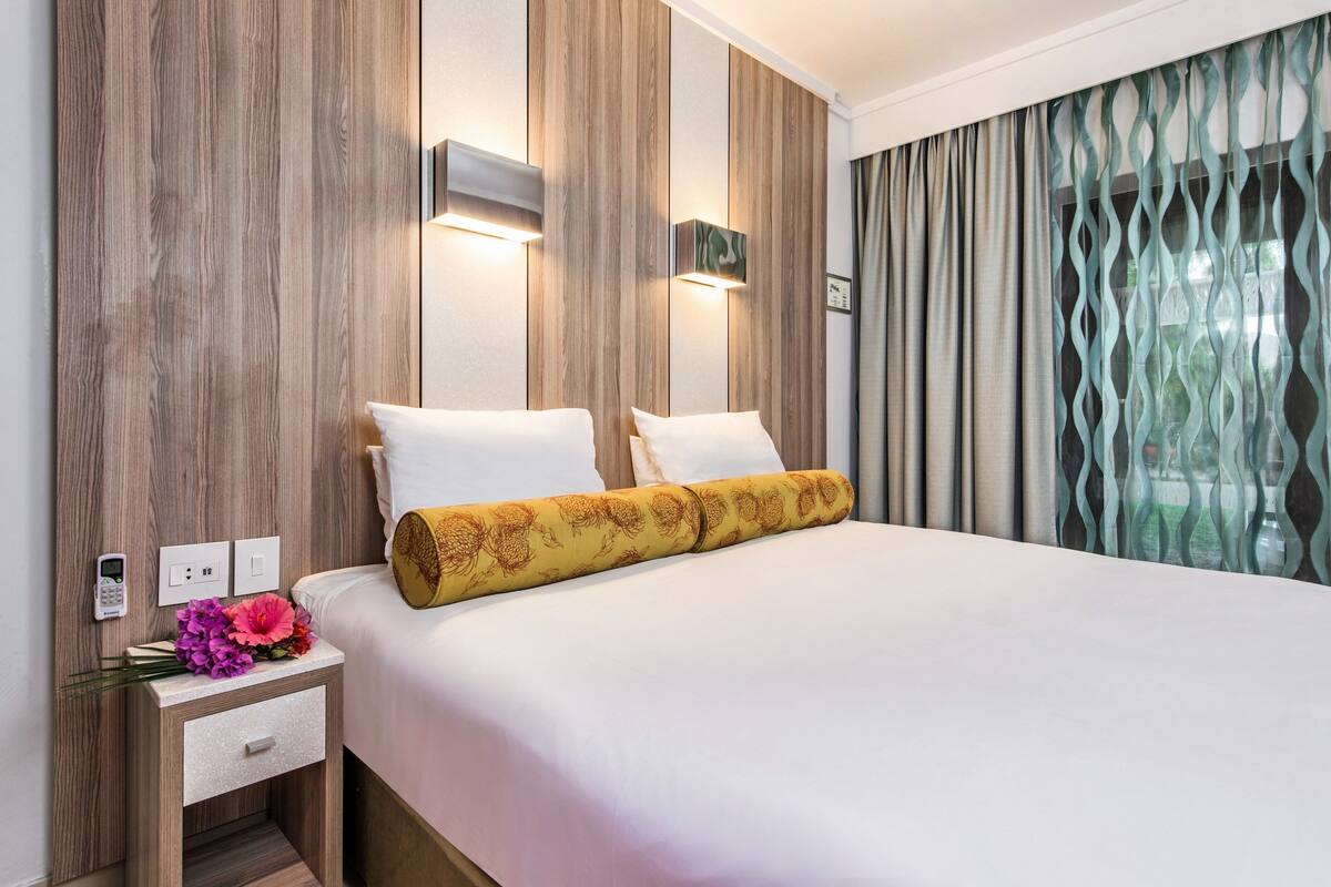 Une chambre d’hôtel avec un grand lit, des murs lambrissés, une table de chevet avec un téléphone et des fleurs, et une vue sur l’extérieur à travers les rideaux.
