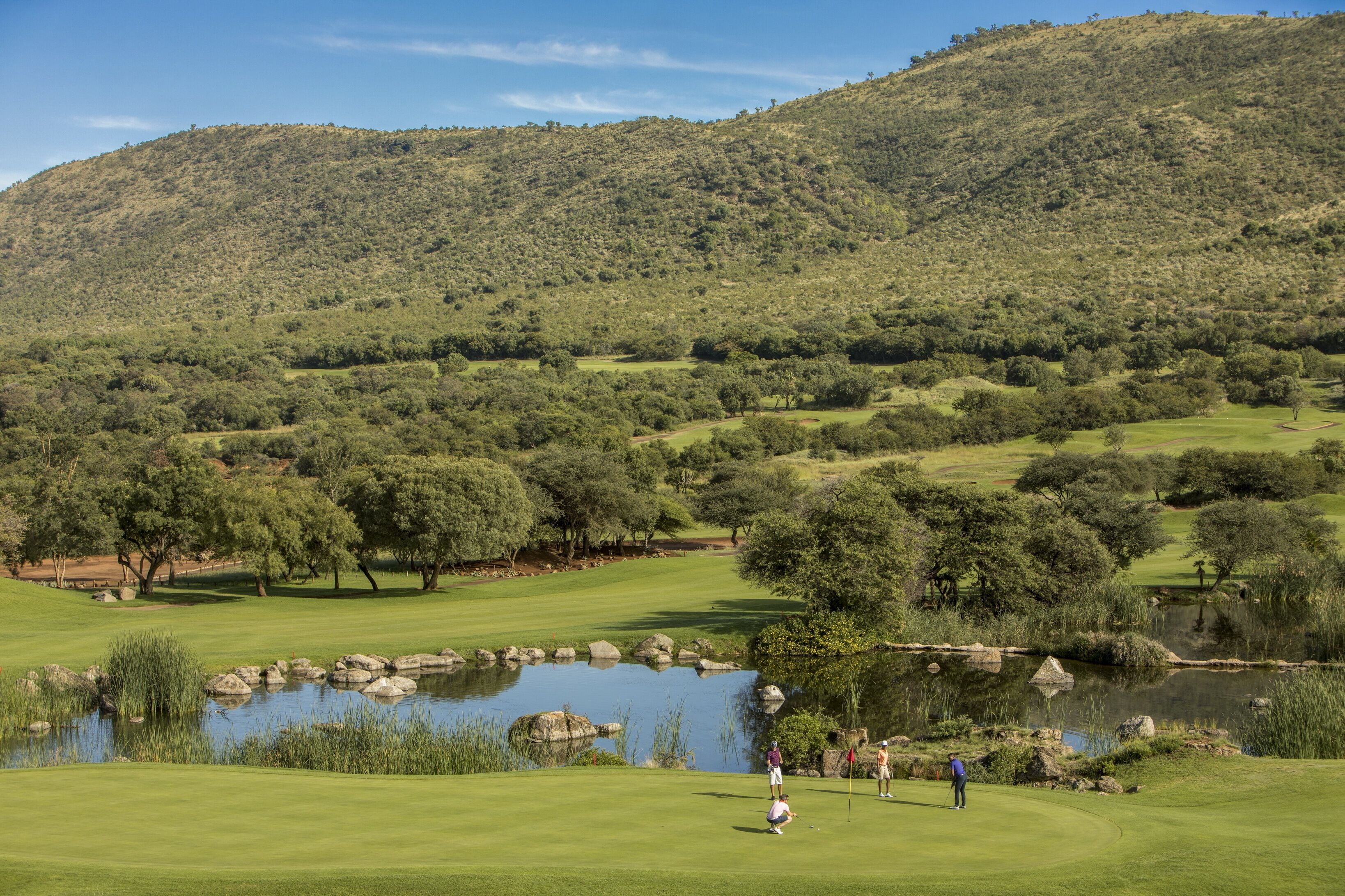 A golf course with a pond, trees, and a hilly background.