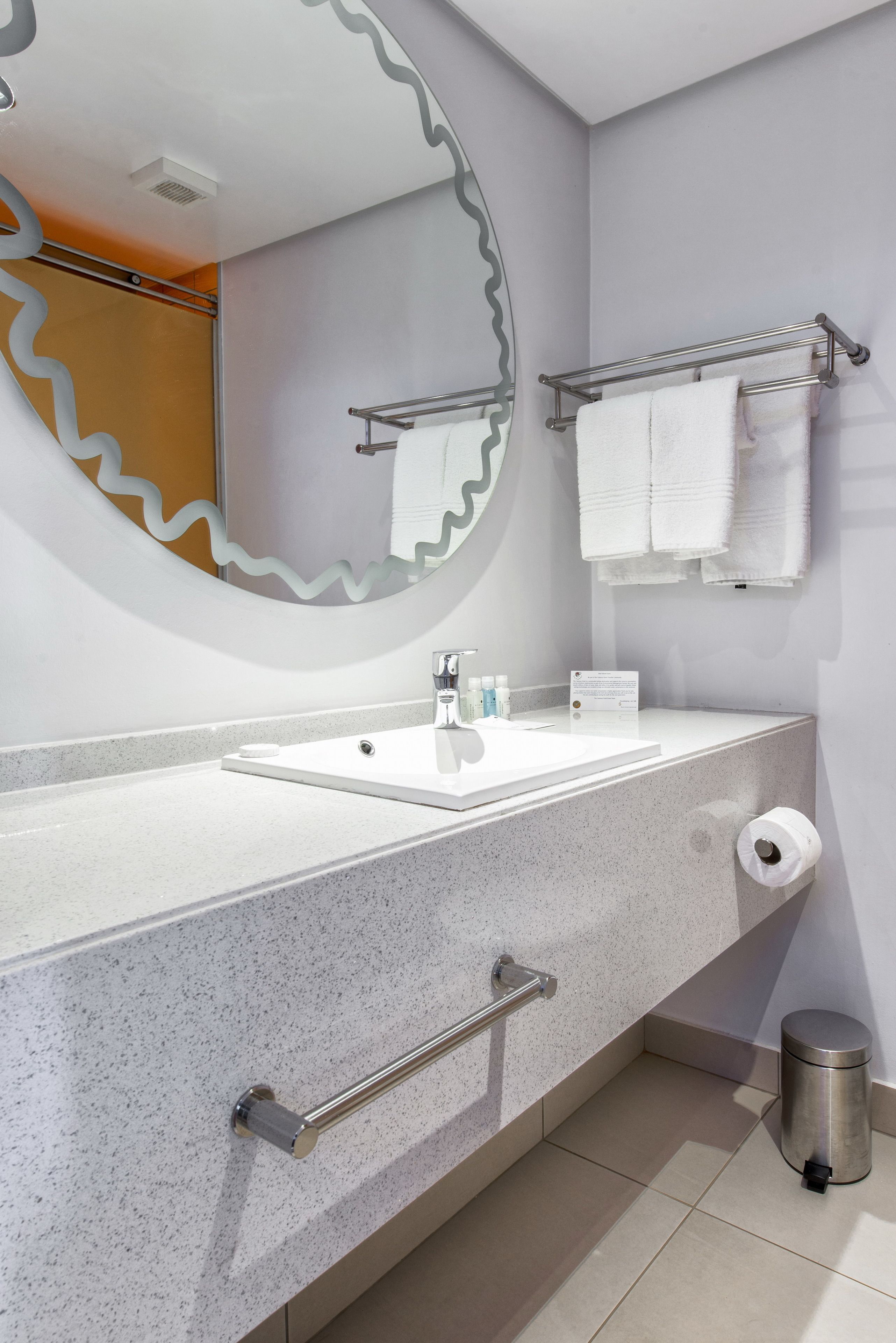 A modern bathroom with a white sink, a large mirror with a wavy edge, a towel rack, and a trash can.