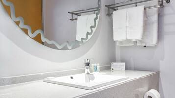 A modern bathroom with a white sink, a large mirror with a wavy edge, a towel rack, and a trash can.