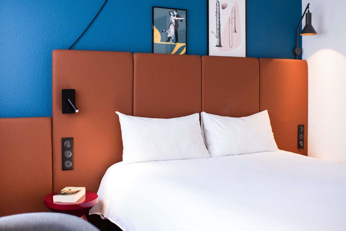 A hotel room with a large bed, a red side table, a book, a framed picture of a clarinet, and a wall-mounted lamp.