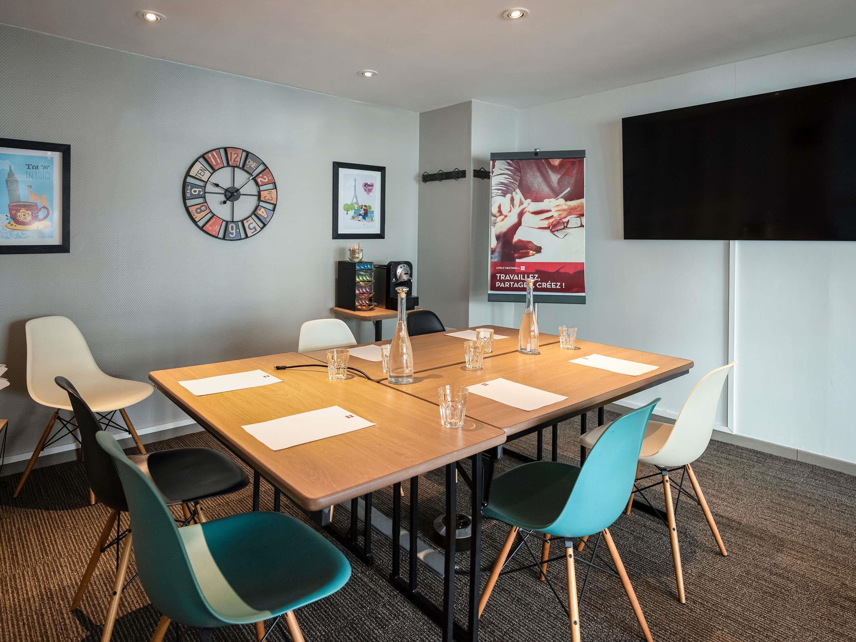 A conference room with a long table, four chairs, a clock, framed pictures, and a TV on the wall.