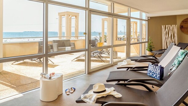 A sunlit room with large windows overlooking a beach, featuring lounge chairs, a small table, and a view of the sea and beach structures.