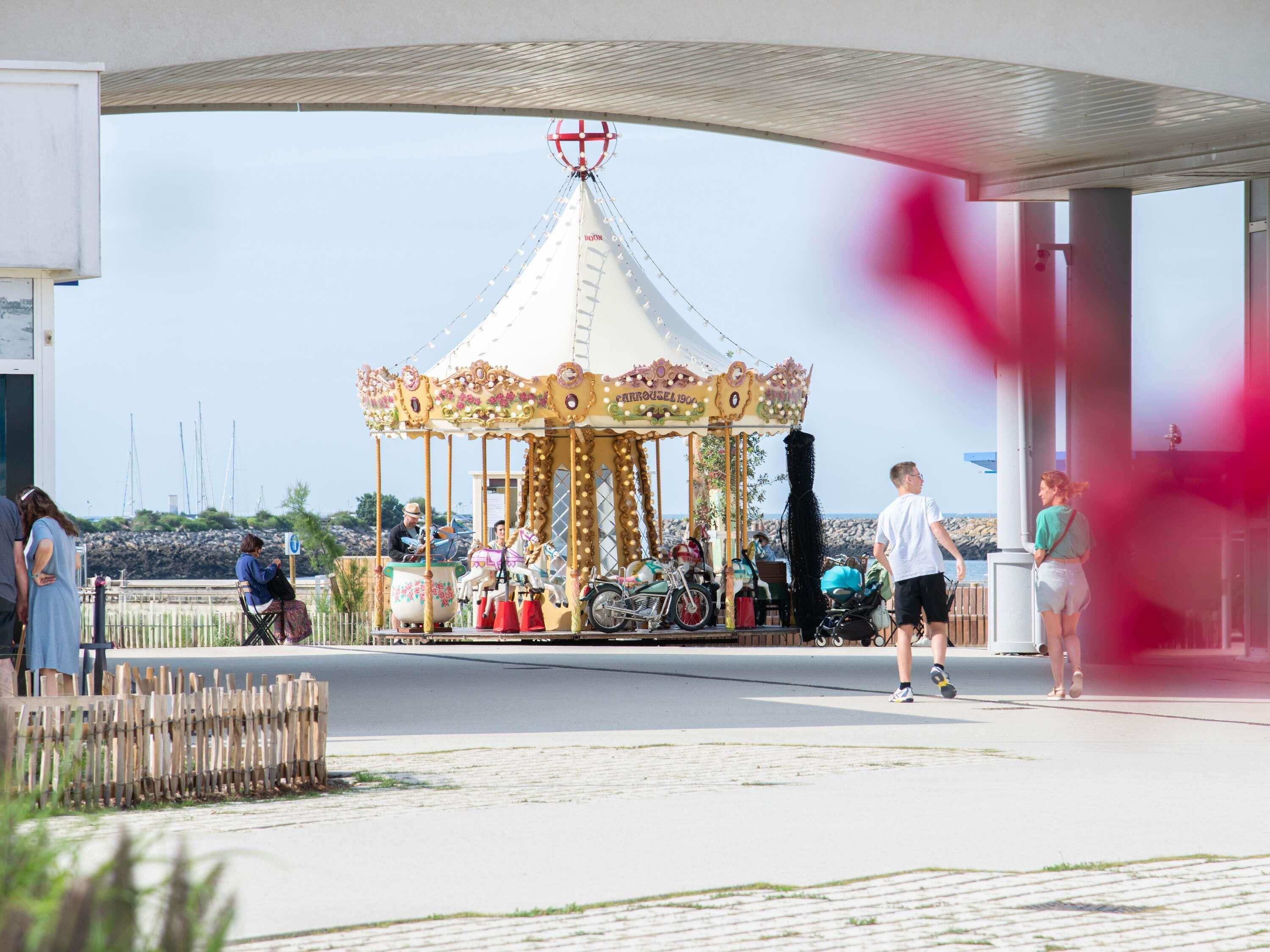 Un carrousel aux couleurs blanc et or, situé dans un espace extérieur doté d’une allée pavée et d’une clôture en bois.