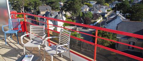 A balcony with white chairs and a magazine on a small table, overlooking a residential area with houses and trees.