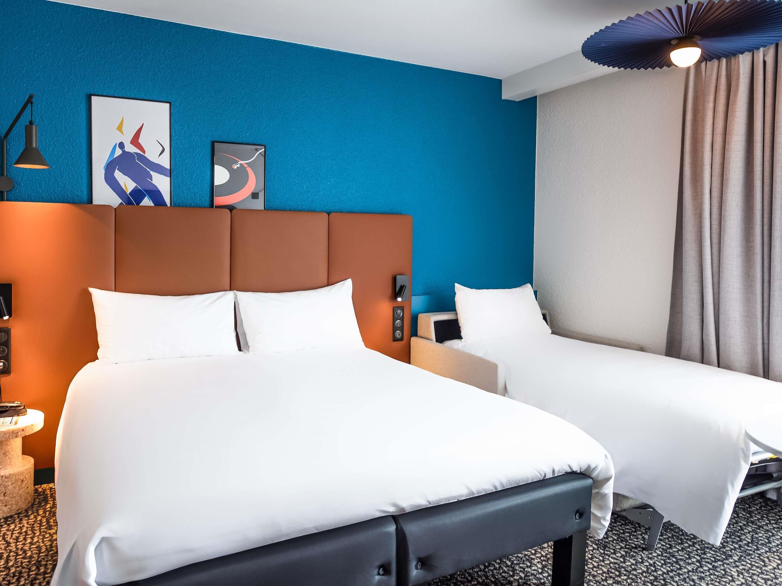 Una habitación de hotel con una cama grande, una cama más pequeña, un cabecero, una mesita de noche, una lámpara y obras de arte en la pared.