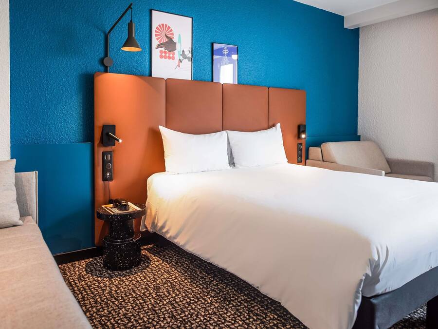 Hotelkamer met een groot bed, een nachtkastje met lamp, een stoel en een muur met kunstwerk.