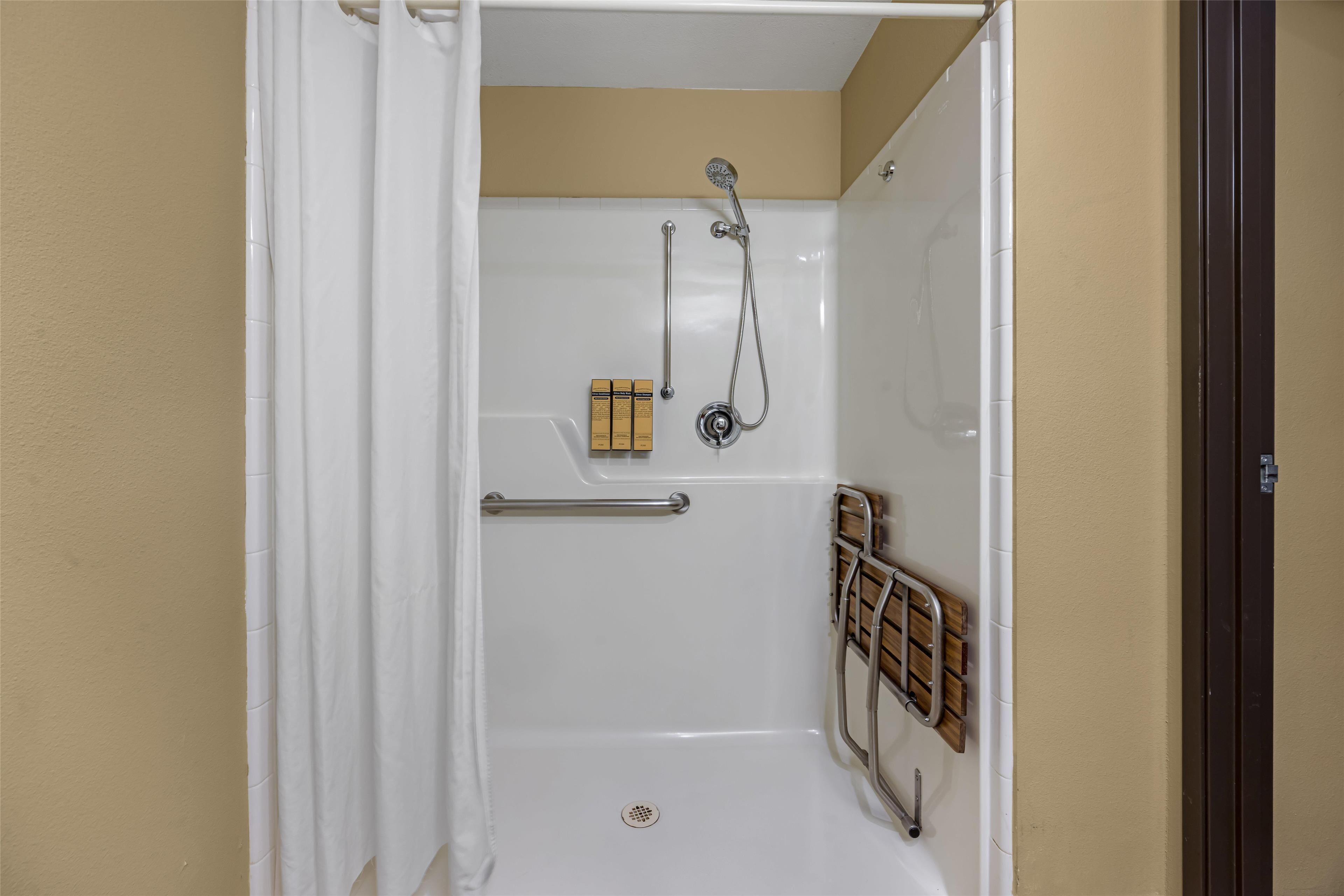 A shower with a white curtain, a handheld showerhead, a handheld soap dispenser, and a wooden shelf with a towel rack.