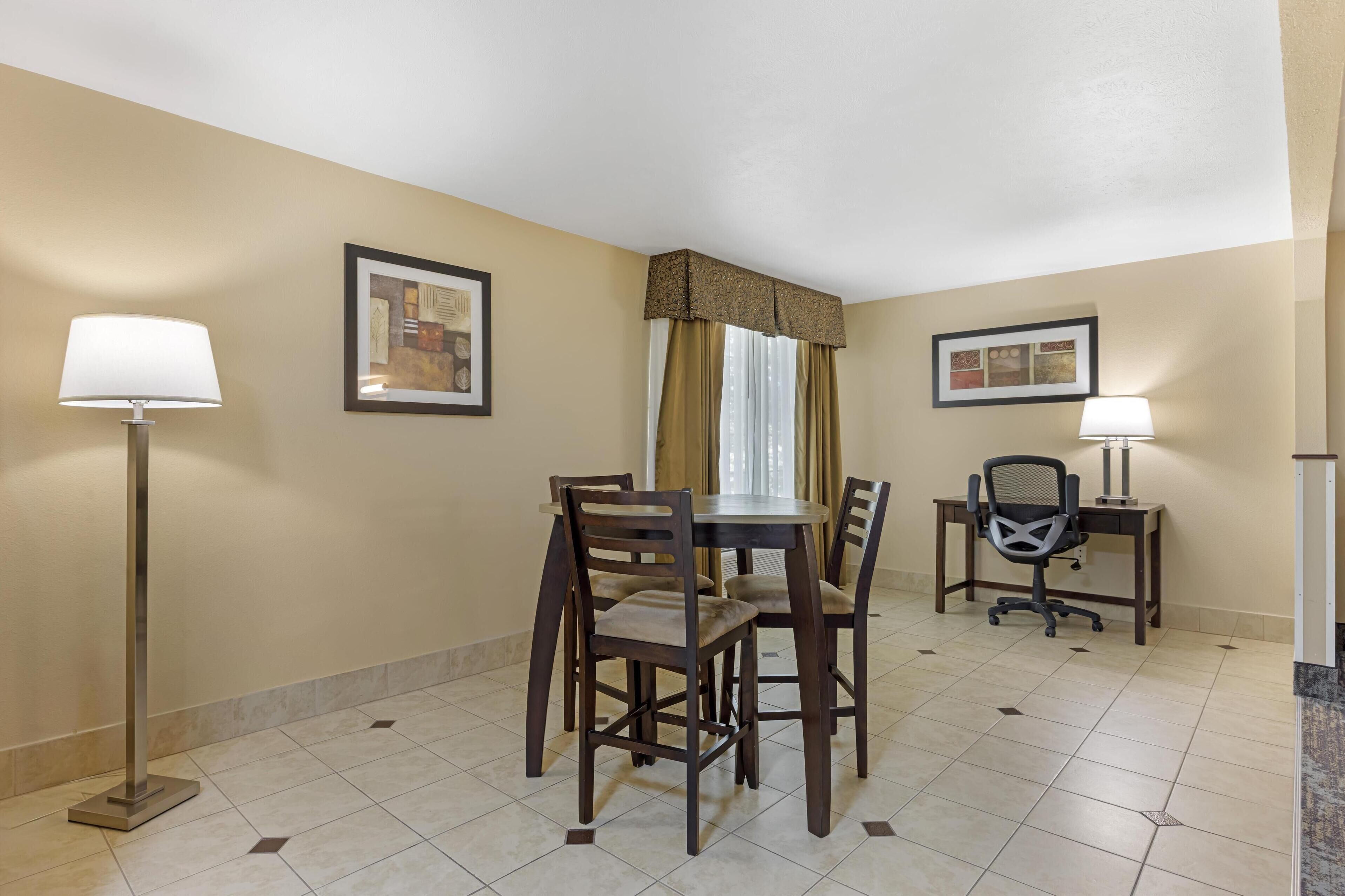 A hotel room with a dining table, four chairs, a desk with a chair, two lamps, and two framed pictures on the walls.