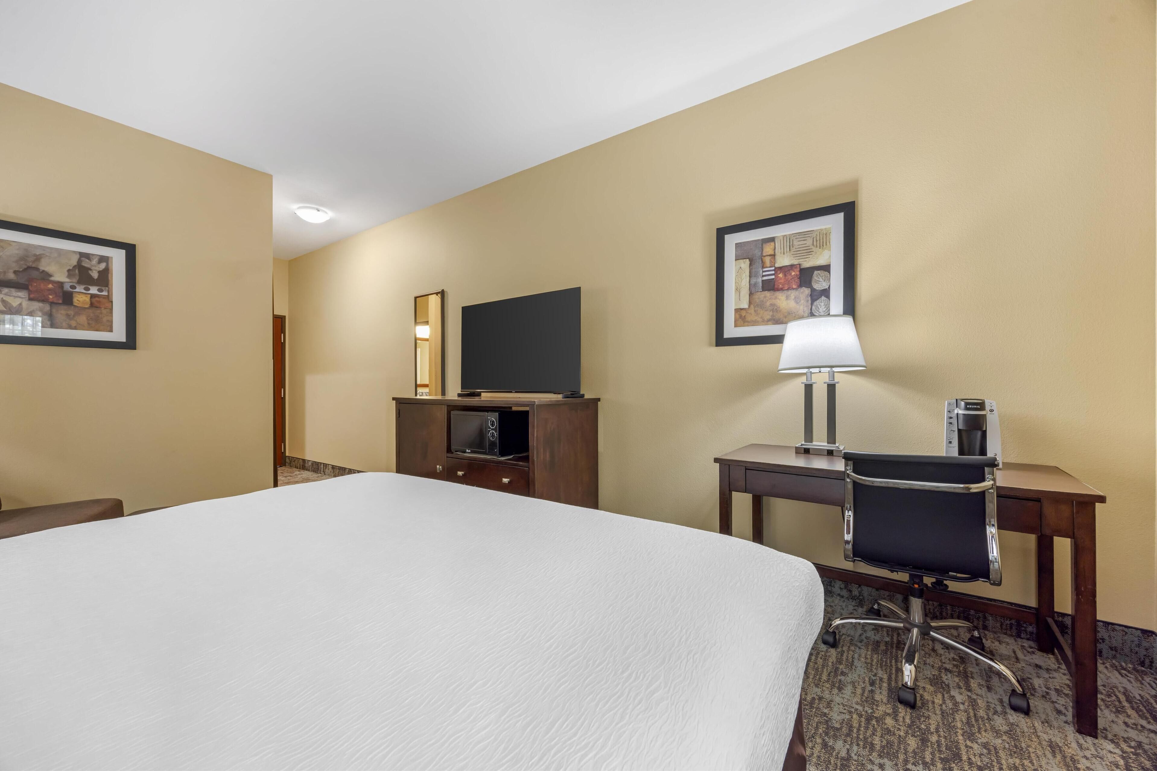 Una habitación de hotel con una cama grande, un escritorio con silla, un televisor de pantalla plana, una lámpara y obras de arte en las paredes.