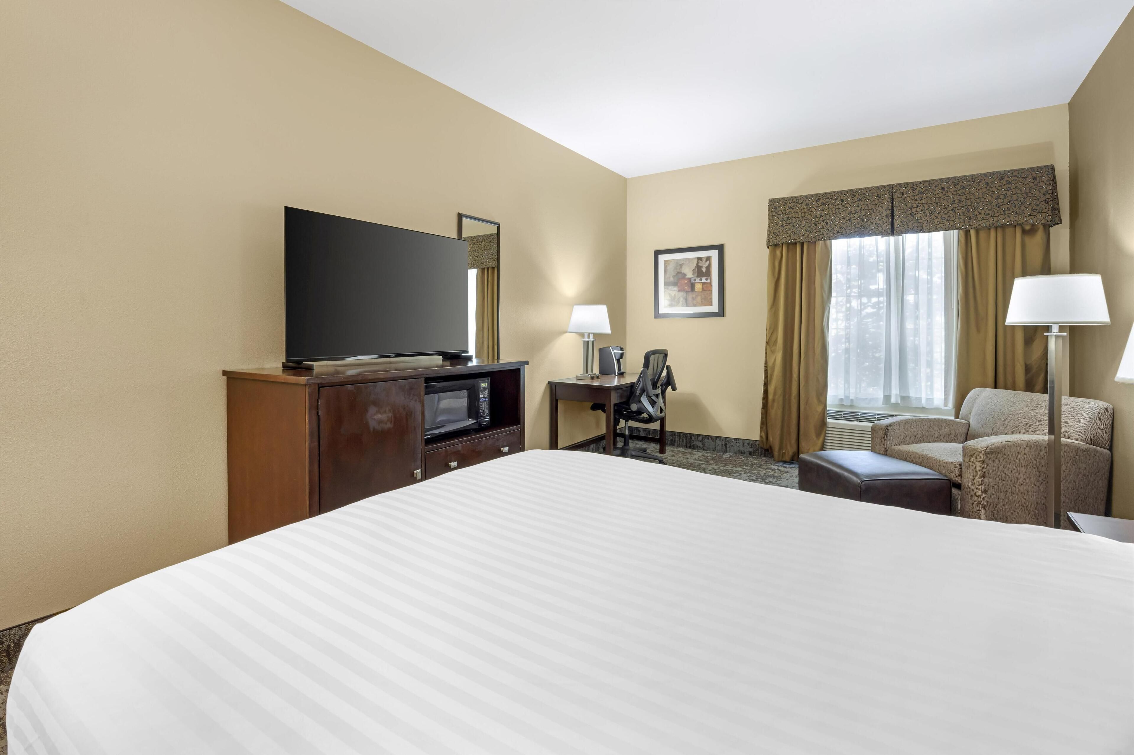 A hotel room with a large bed, a flat-screen TV, a desk with a chair, and a window with curtains.