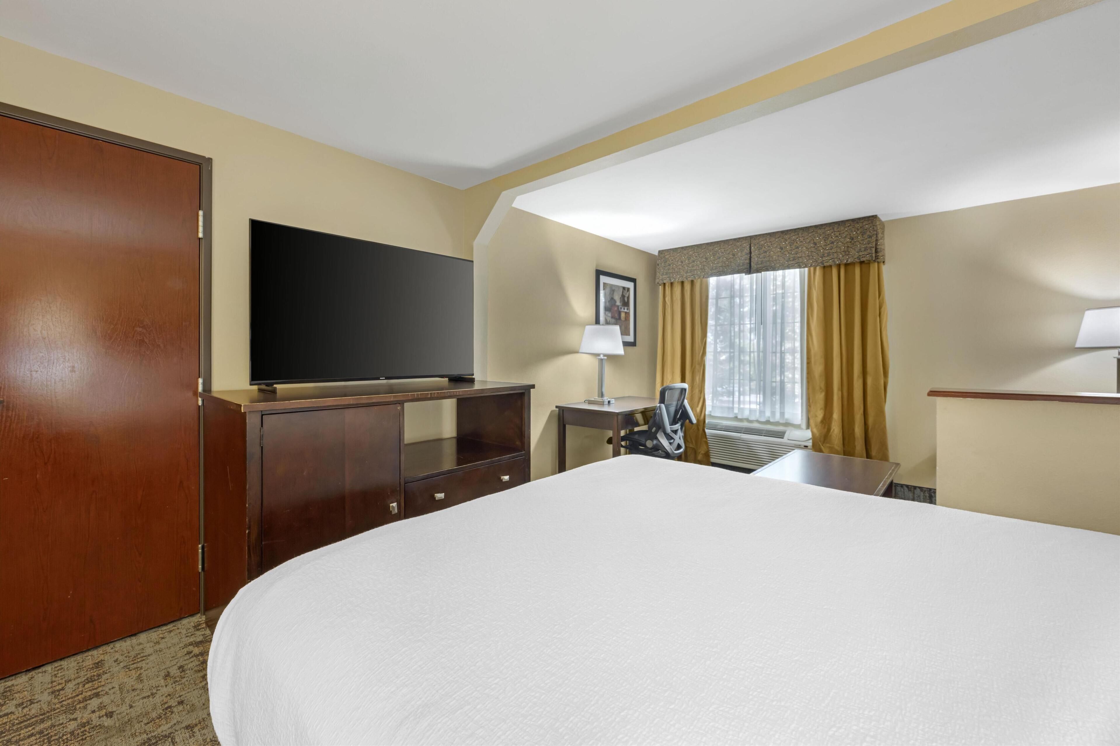 A hotel room with a large bed, a desk with a chair, a television, and a window with curtains.